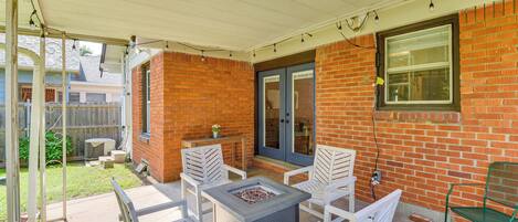 A patio with a fire pit table and white chairs, a brick wall, and a covered area with string lights.