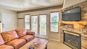 A living room with a leather sofa, a coffee table with a book and remote, a fireplace, a TV mounted on a stone wall, and a door with a white frame. - Cozy Eden Condo Retreat < 6 Mi to Ski Resort! (Eden)