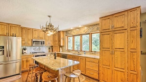 A kitchen with wooden cabinets, a stainless steel refrigerator, a microwave, and a large island with bar stools. - Tranquil Riverside Home w/ Wraparound Deck & Views (Baldwin)