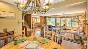 A spacious living area with a wooden dining table set, a chandelier, and large windows offering a view of the outdoors. - Tranquil Riverside Home w/ Wraparound Deck & Views (Baldwin)