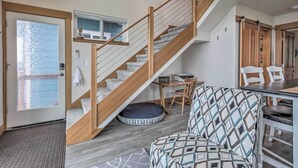 Una escalera con barandales de madera que conduce a un segundo piso, un comedor con mesa y sillas, y vista al exterior a través de un ventanal.