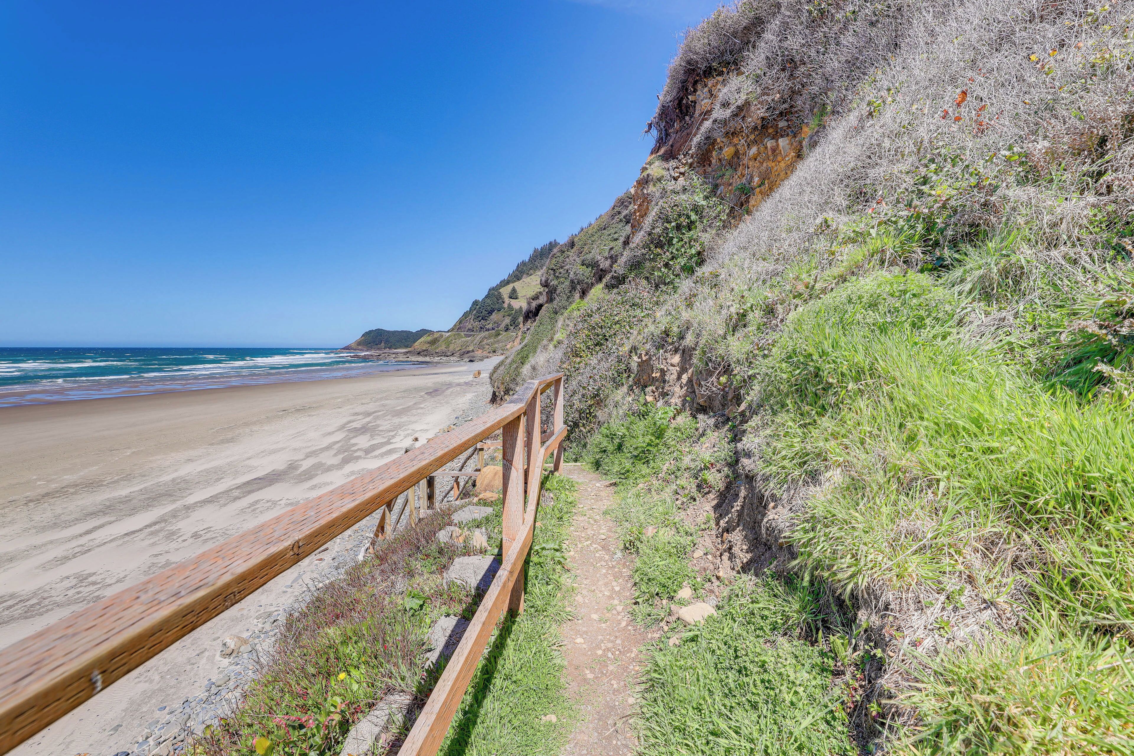 Un sentier côtier bordé d’une rambarde en bois, qui mène à une plage où l’on peut admirer les vagues et un ciel bleu dégagé.