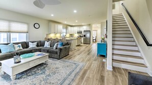 A spacious living room with a gray sectional sofa, a white coffee table, and a blue patterned rug. A kitchen with white cabinets and a stainless steel refrigerator is visible in the background. - Sunny Brigantine Townhome: Walk to Beach (Brigantine)