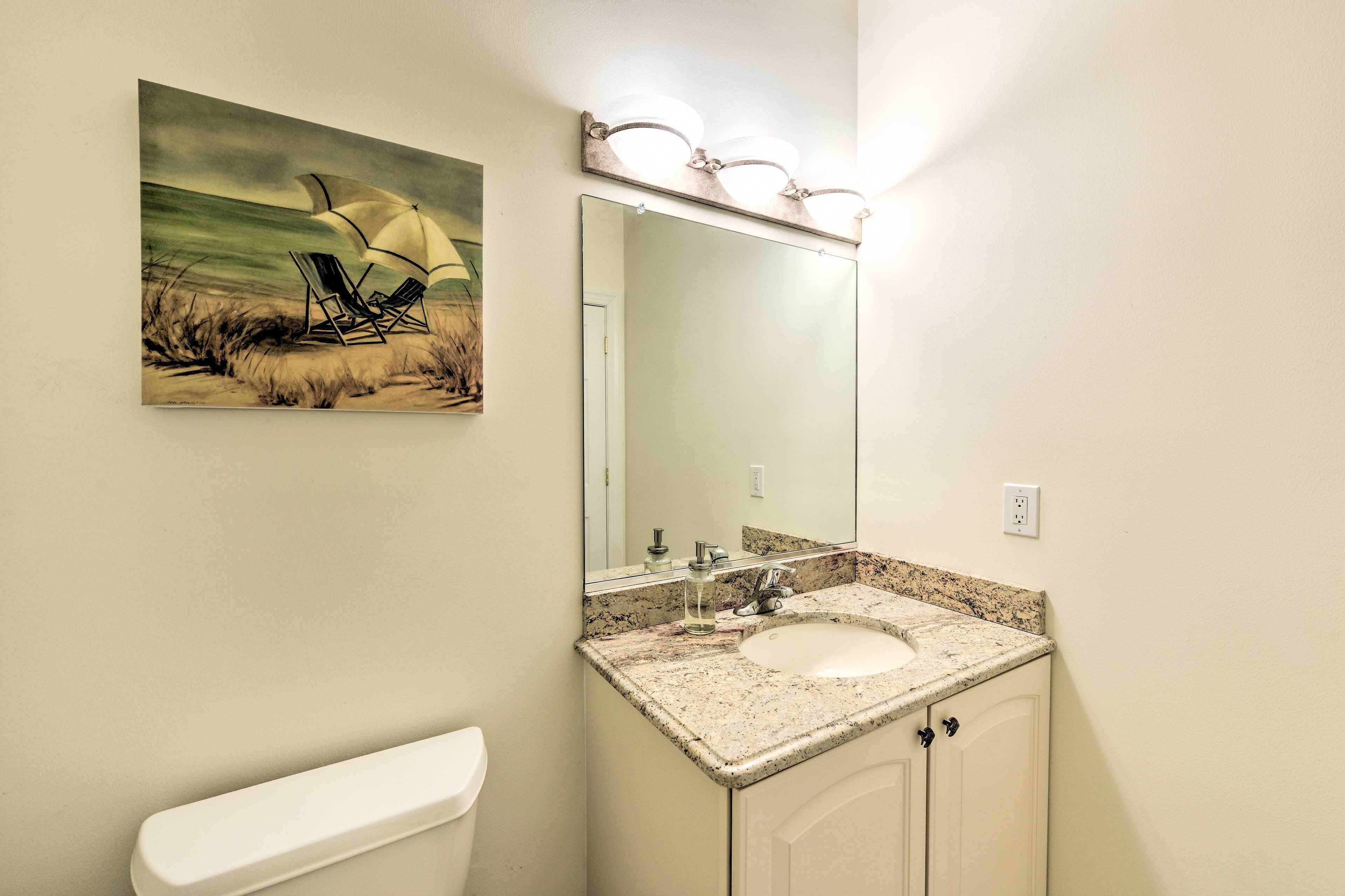 Une salle de bain avec un lavabo, un miroir et des toilettes. Au-dessus du miroir, on peut voir un tableau représentant une scène de plage.