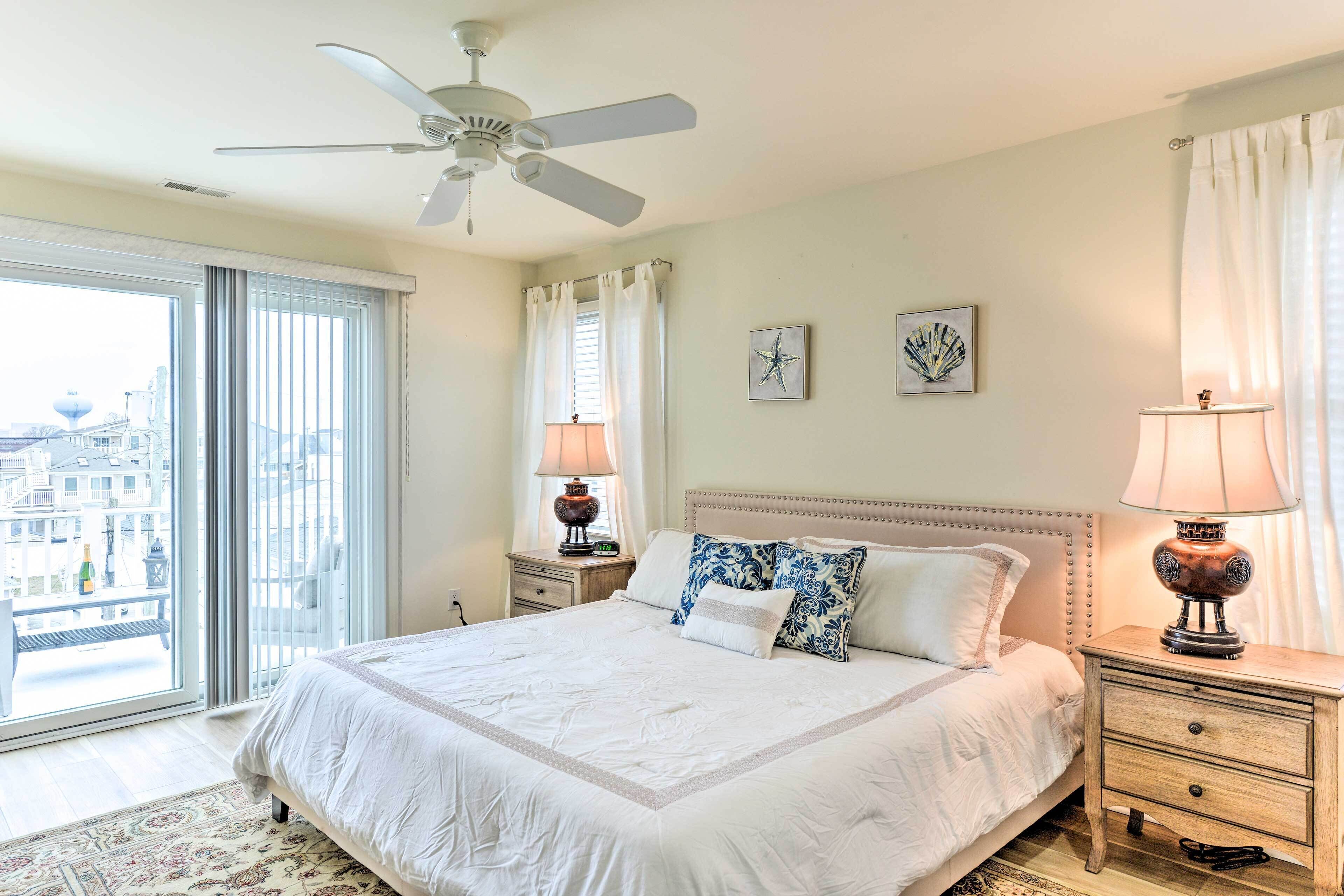 A bedroom with a large bed, two bedside tables with lamps, a ceiling fan, and a view of the city through large windows.