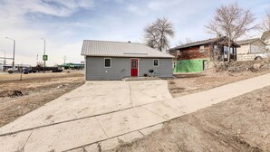 A small, single-story house with a red door and a metal roof, surrounded by a concrete driveway and a dirt path. - Pierre Vacation Rental w/ Grill & Fenced Yard (Pierre)