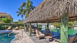 A pool area with lounge chairs, a thatched roof structure, and a view of a house in the background. - Marathon Vacation Rental With Pool Access! (Marathon)