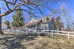 A two-story house with a white fence, a large tree, and a clear blue sky. - Lakefront Camdenton Home w/ Fire Pit & Grill! (Camdenton)