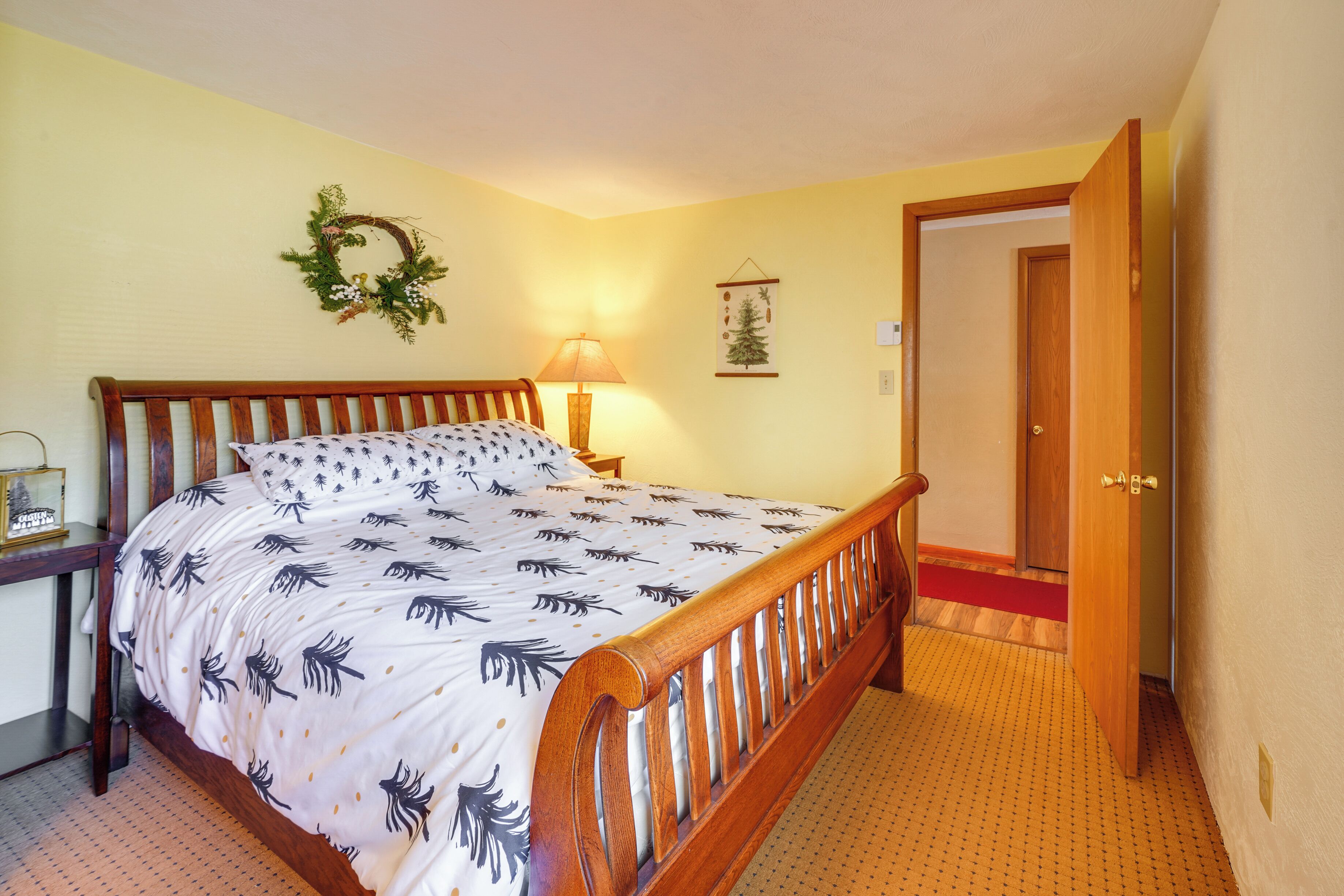Een slaapkamer met een houten bed, een nachtkastje, een lamp, een aan de muur bevestigd krans en een deur naar een andere kamer.