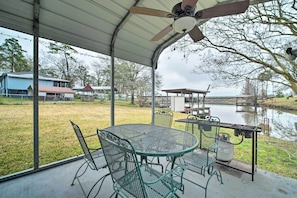 Um alpendre coberto com mesa e cadeiras, churrasqueira a gás, e vista para um corpo d'água e edifícios ao fundo.