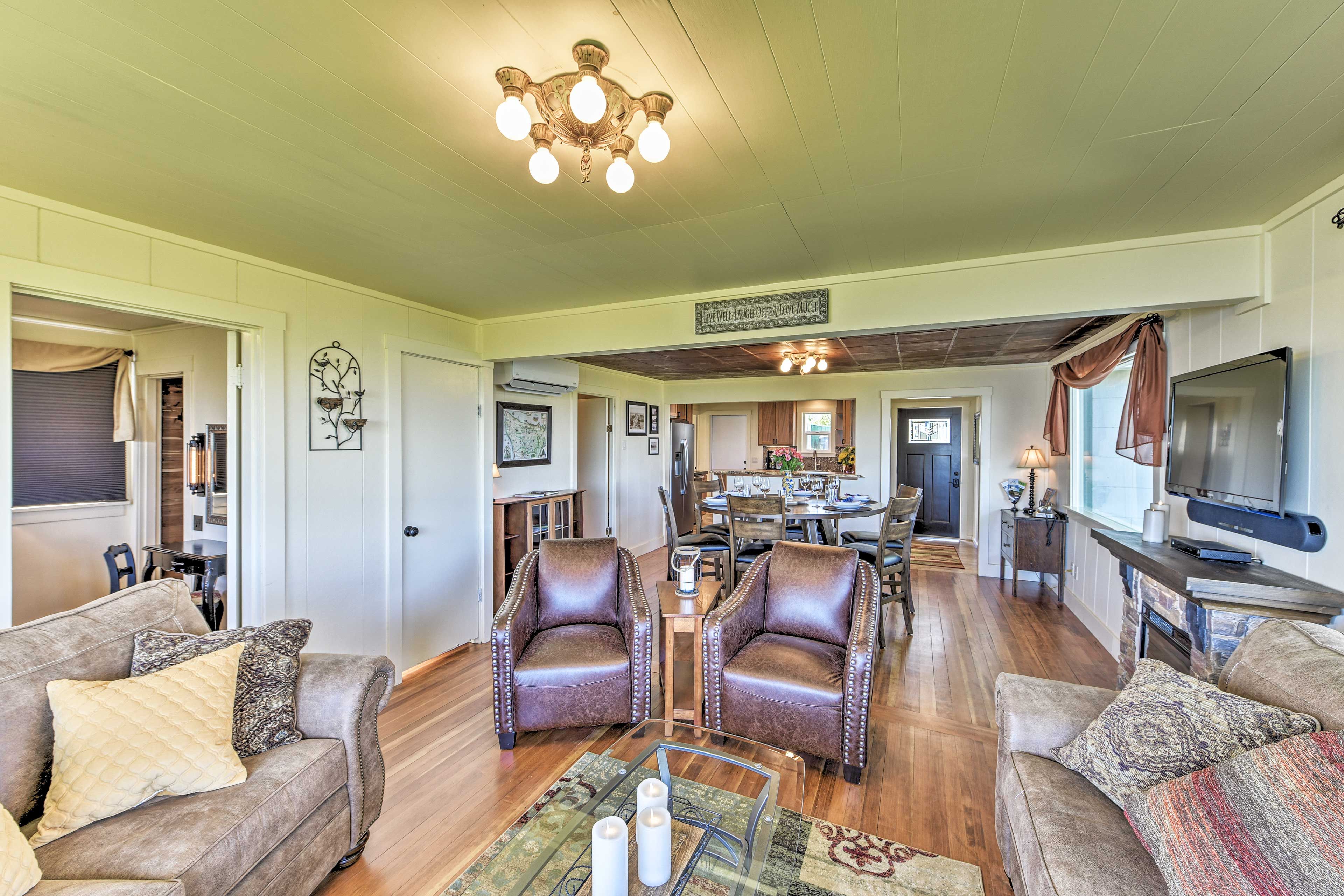 A spacious living room with leather chairs, a glass coffee table, and a television. There is a dining area in the background with a chandelier.