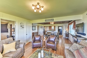 A spacious living room with leather chairs, a glass coffee table, and a television. There is a dining area in the background with a chandelier. - Waterfront Home ~ 6 Mi to Olympic National Park! (Port Angeles)