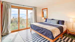 A bedroom with a large bed, a view through the window, and a bedside table with a lamp. - Serene Jonesport Escape: Deck w/ Water Views! (Jonesport)