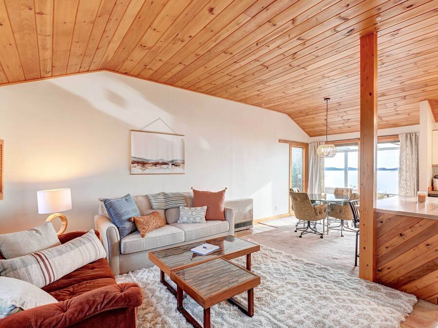 A cozy living room with a wooden ceiling, a sofa set, a coffee table, and a view of the kitchen and dining area.