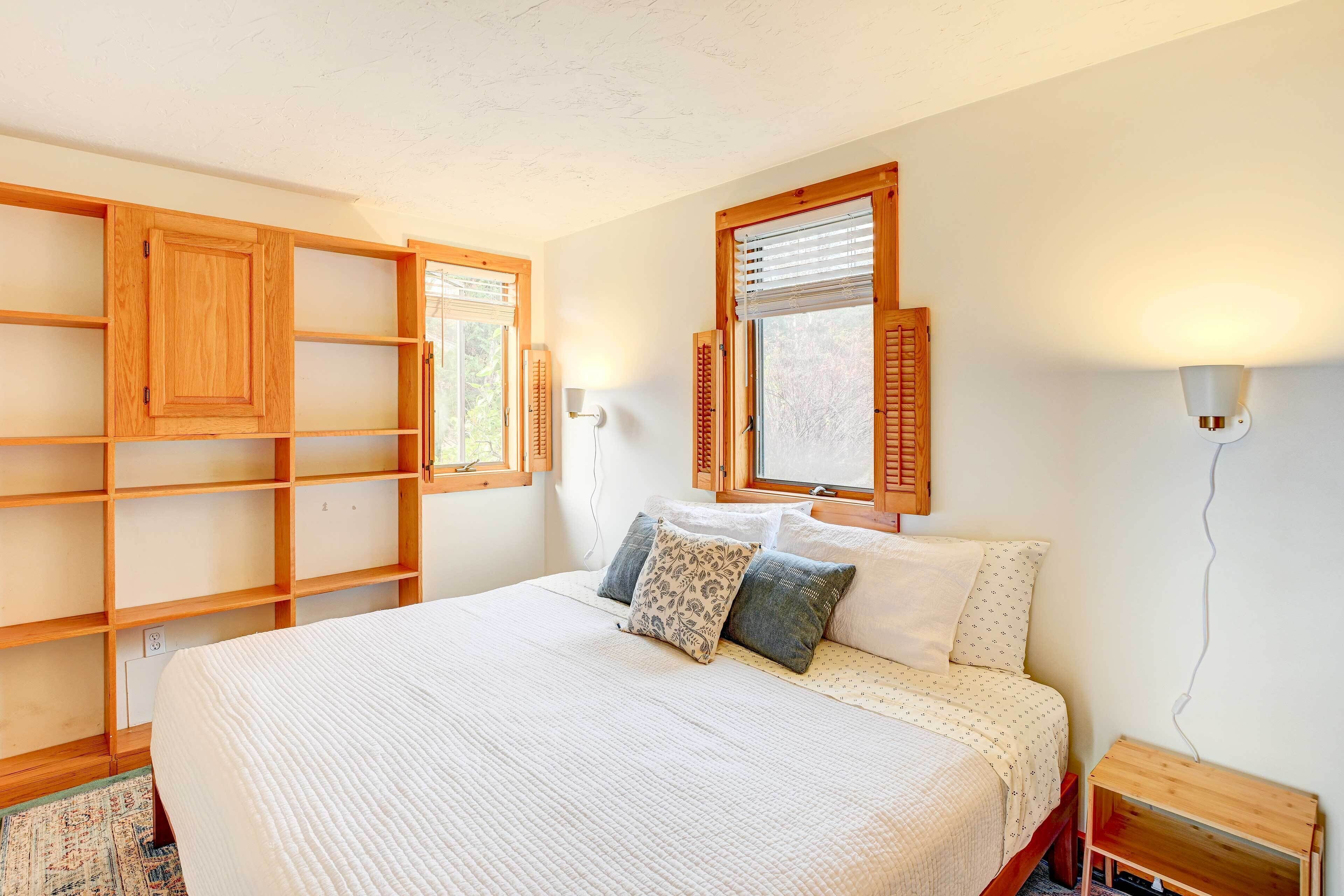 A bedroom with a large bed, wooden shelves, two windows with shutters, and a bedside table.