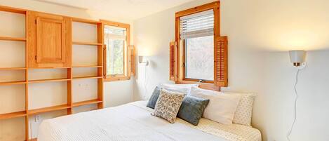 A bedroom with a large bed, wooden shelves, two windows with shutters, and a bedside table.