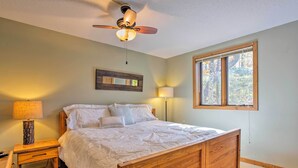 Une chambre avec un lit en bois, un ventilateur de plafond, une table de chevet avec une lampe, une commode et une fenêtre donnant sur des arbres.