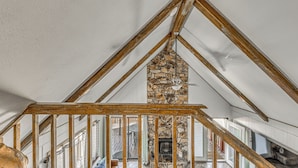 A high-ceilinged room with exposed wooden beams, a stone fireplace, and a view of an outdoor patio.