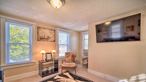 A living room with a large window, a sofa, a chair, a rug, and a TV mounted on the wall. - Family-friendly Wildwood Apt: Bike to Beach! (Wildwood)