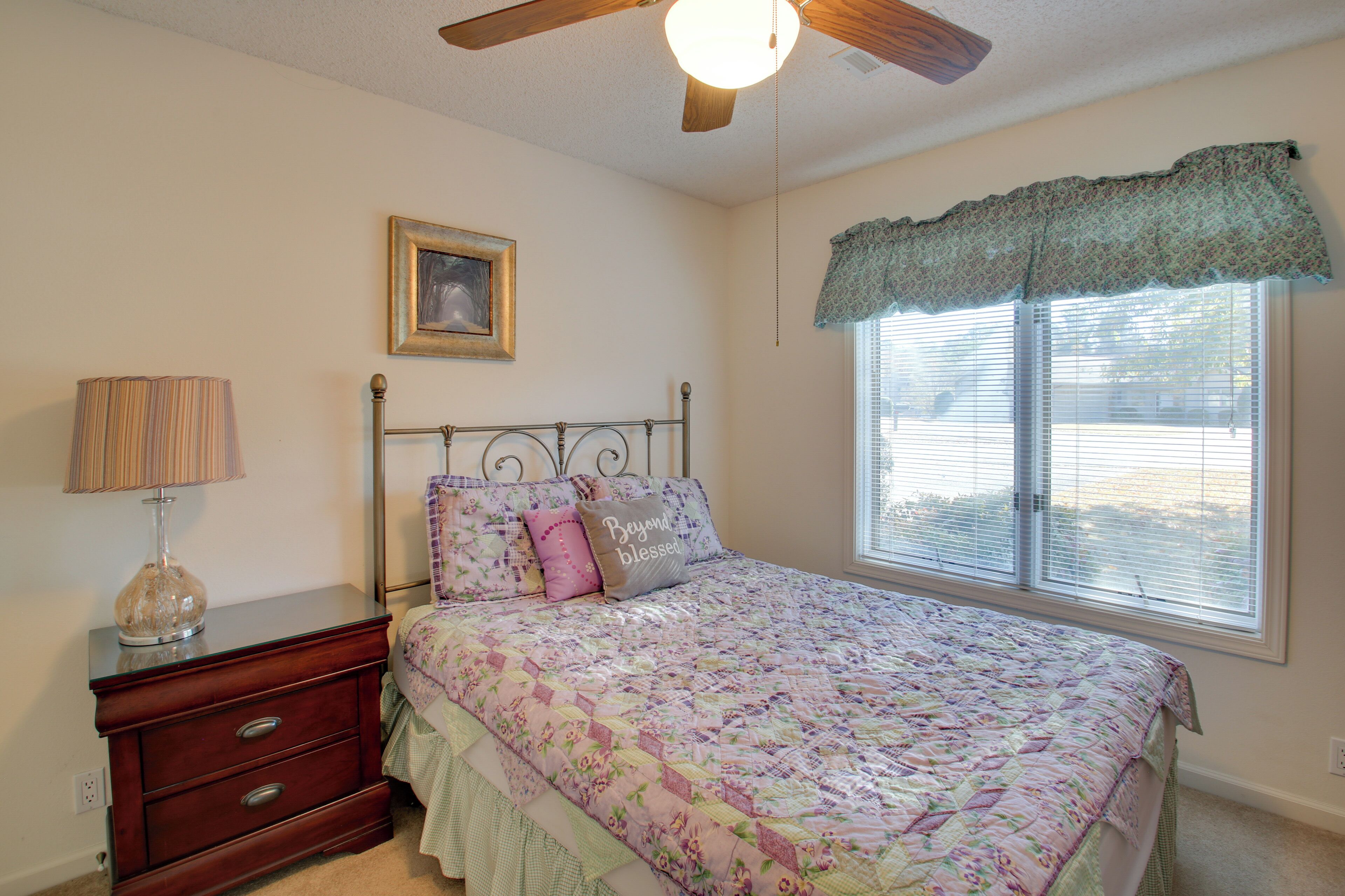 A bedroom with a bed, a nightstand, a lamp, a ceiling fan, a window with blinds, and a framed picture on the wall.