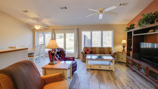 A spacious living room with a sofa, armchair, coffee table, and a television. There are ceiling fans, a dining area with chairs, and a large window providing natural light. - Lake Balboa Access: Home w/ Dock & Yard (Hot Springs Village)
