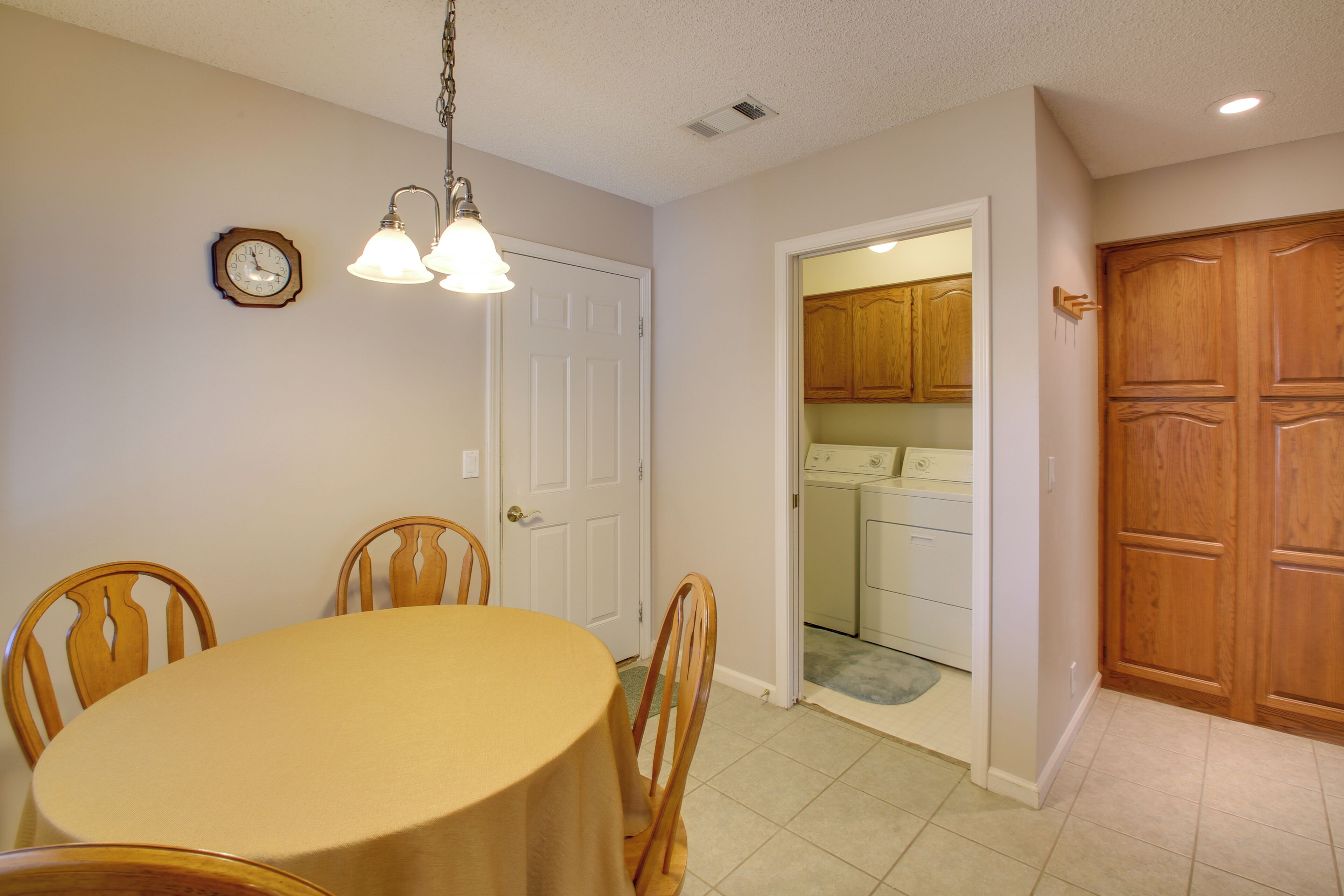 A dining area with a round table and wooden chairs, a clock on the wall, and a wooden door leading to a laundry room.
