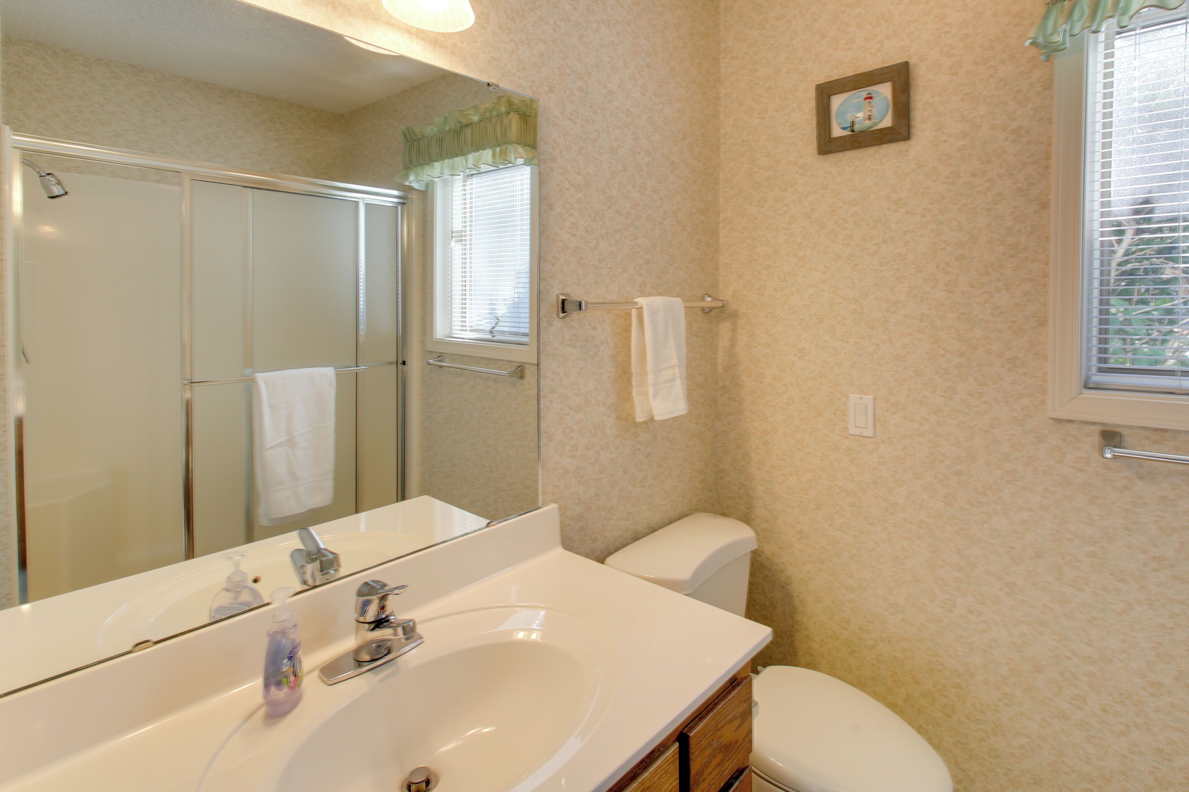 A bathroom with a white sink, mirror, and toilet. There is a shower door and a window with blinds.