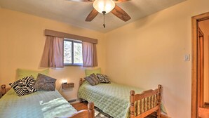 Quarto com duas camas de madeira, um ventilador de teto, uma janela com cortinas e uma mesa de cabeceira com abajur.