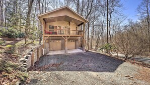 Une cabane à deux étages dotée d’un balcon, d’un garage et d’une allée en gravier, le tout entouré d’arbres.