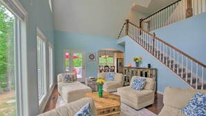 A spacious living room with a high ceiling, a staircase, and large windows.