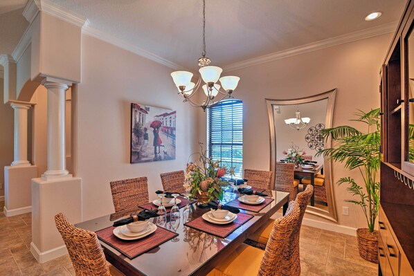 A dining room with a long table set for six, surrounded by wicker chairs, a chandelier, and a large window with blinds.