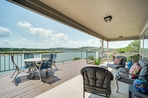 Un balcón con mesa y sillas, un sofá con cojines, y vistas a un lago y colinas.