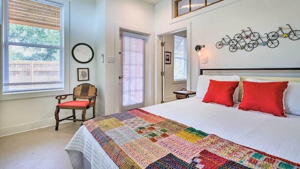 A bedroom with a large bed, two red pillows, a colorful quilt, a wooden chair with a red cushion, a wall-mounted bicycle decoration, and a window with blinds. - Walk to Beach From Chic Old Town Apartment w/ Yard (Bay St Louis)
