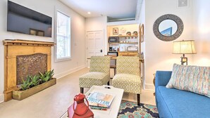 A living room with a fireplace, two patterned armchairs, a blue sofa, a flat-screen TV, and a kitchen area in the background. - Walk to Beach From Chic Old Town Apartment w/ Yard (Bay St Louis)