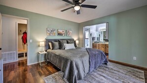 A bedroom with a bed, a ceiling fan, a nightstand, and a door leading to a bathroom. - Spacious Bella Vista Home ~ 5 Mi to Lake! (Bella Vista)