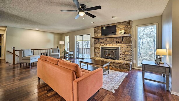 A spacious living room with hardwood flooring, a stone fireplace, a ceiling fan, and a large window providing natural light. - Spacious Bella Vista Home ~ 5 Mi to Lake! (Bella Vista)
