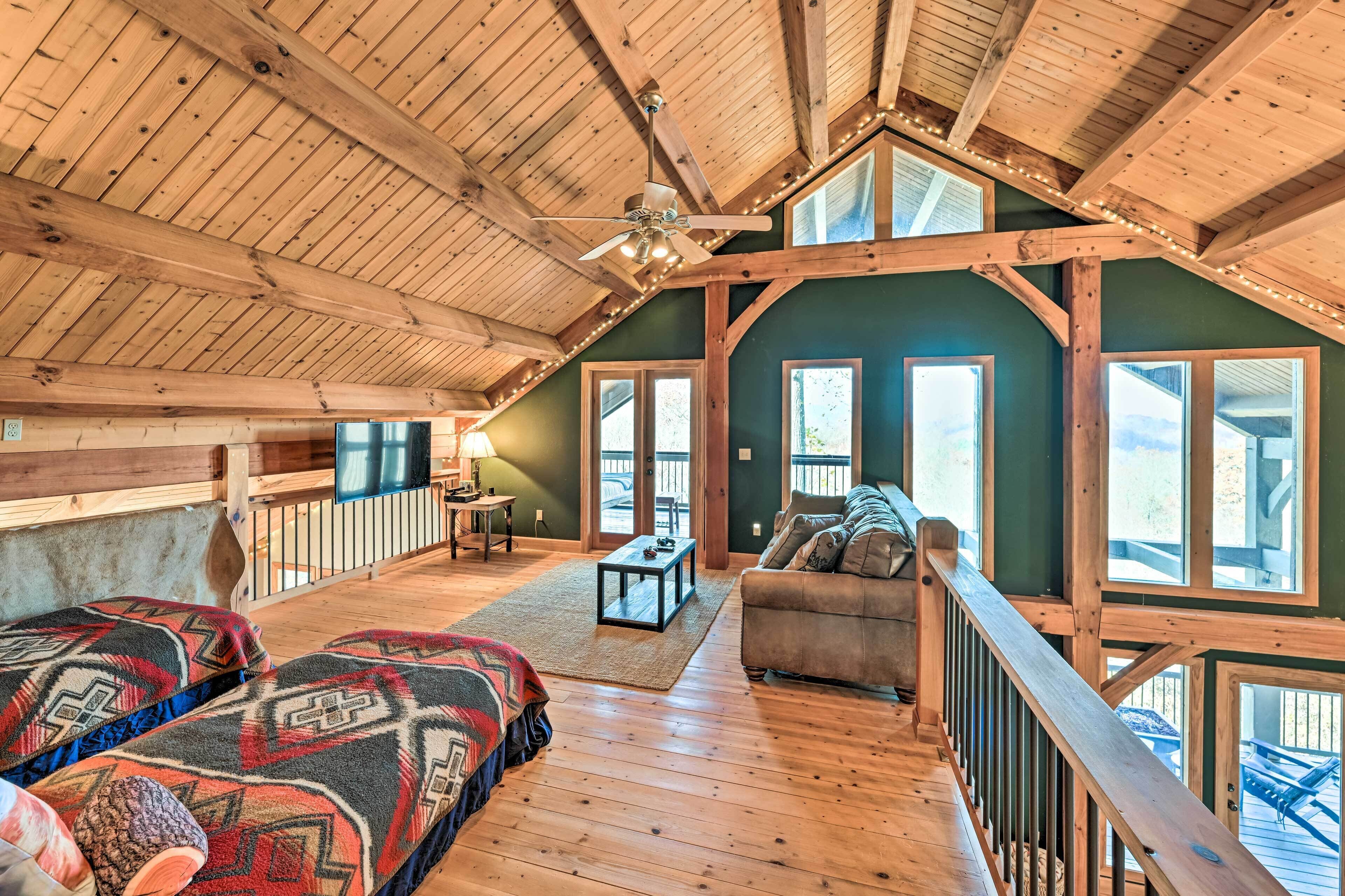 A spacious loft bedroom with a high ceiling, wooden beams, a large bed, a sofa, and a view of the outdoors.
