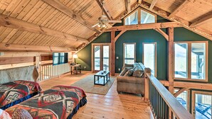 A spacious loft bedroom with a high ceiling, wooden beams, a large bed, a sofa, and a view of the outdoors.