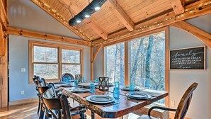 A dining area with a wooden table set for four, surrounded by chairs, large windows with a view of trees, and a sign on the wall.