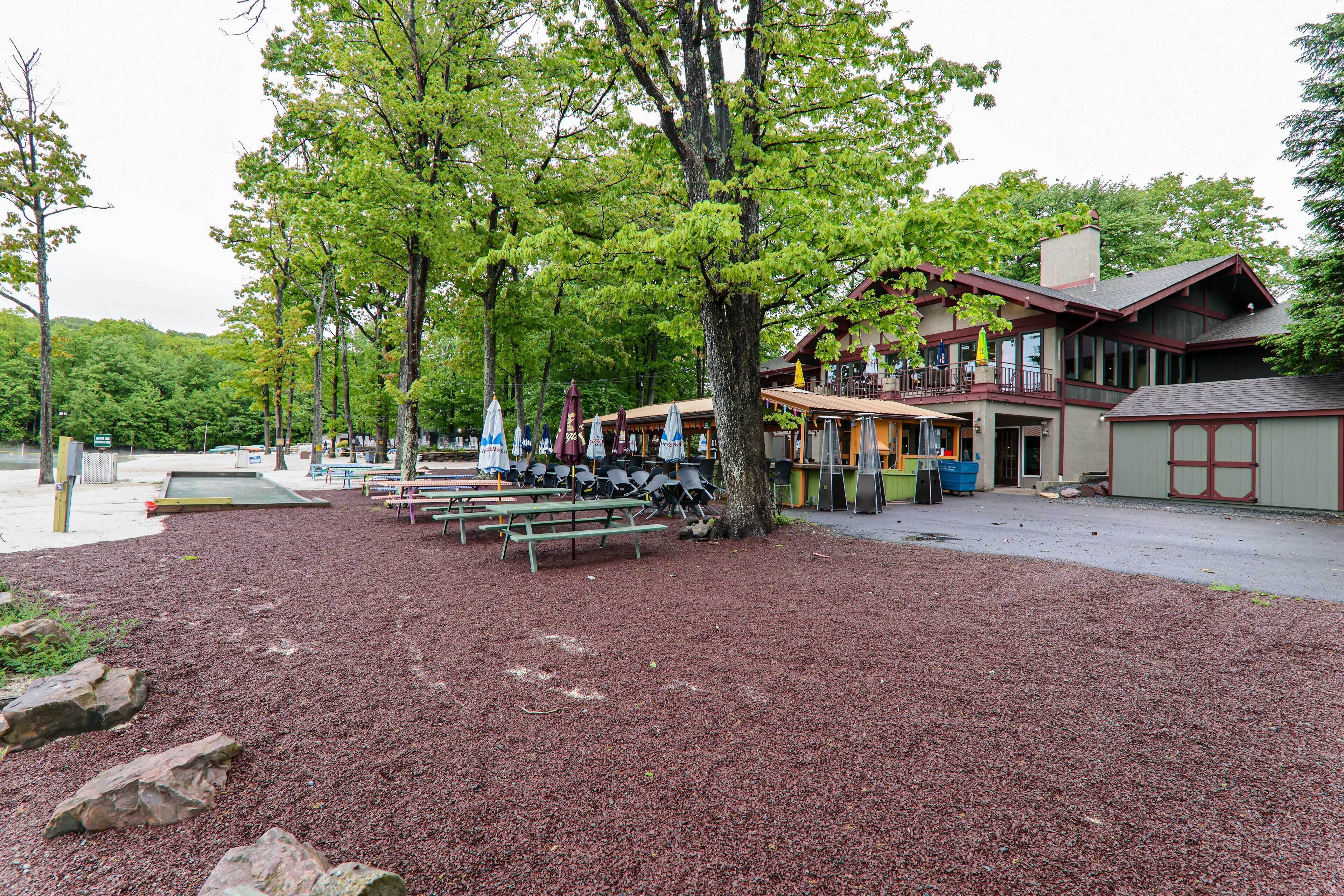 Un espace aménagé en parc, avec des tables de pique-nique, des bancs et des parasols, entouré d’arbres et bordé d’un bâtiment doté d’une terrasse.
