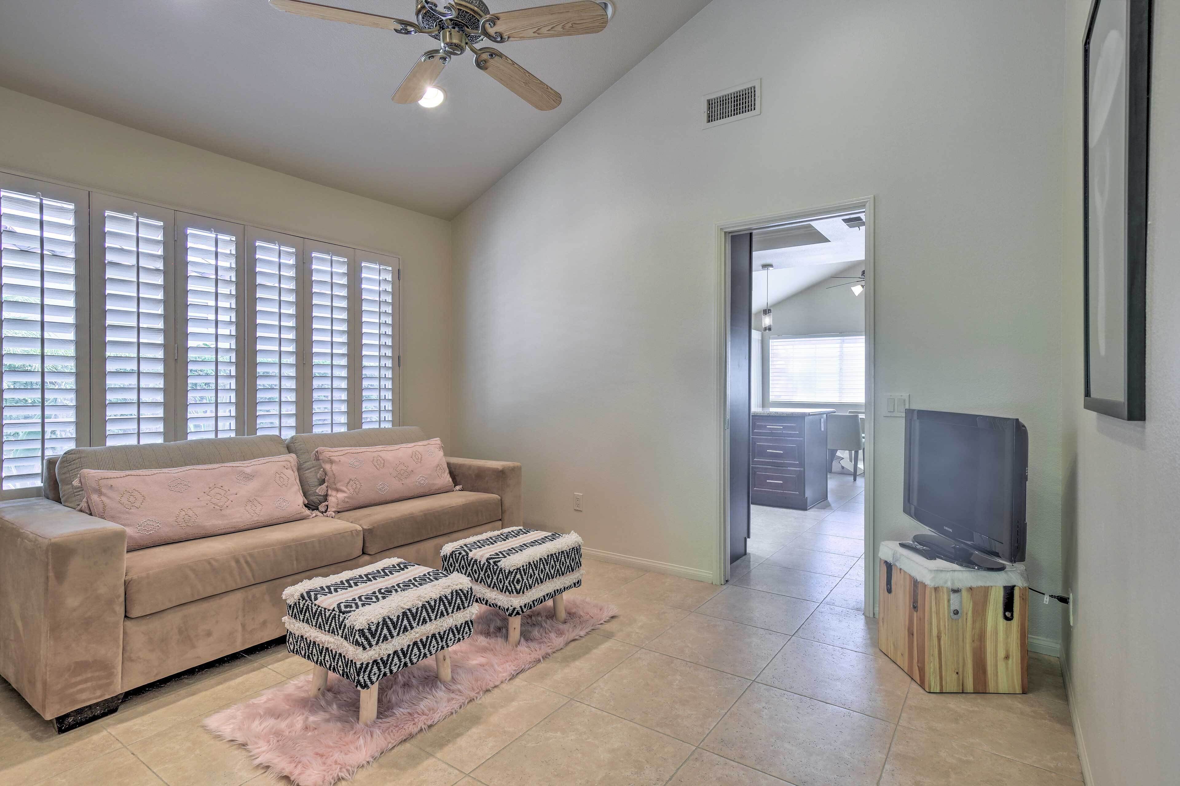 A living room with a beige sofa, two patterned ottomans, a television on a wooden stand, and a ceiling fan.