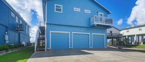 A two-story residential building with a blue exterior, a balcony on the second floor, and three garage doors.
