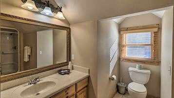 Une salle de bain avec un grand miroir, un lavabo, des meubles en bois, des toilettes et une fenêtre avec des stores.