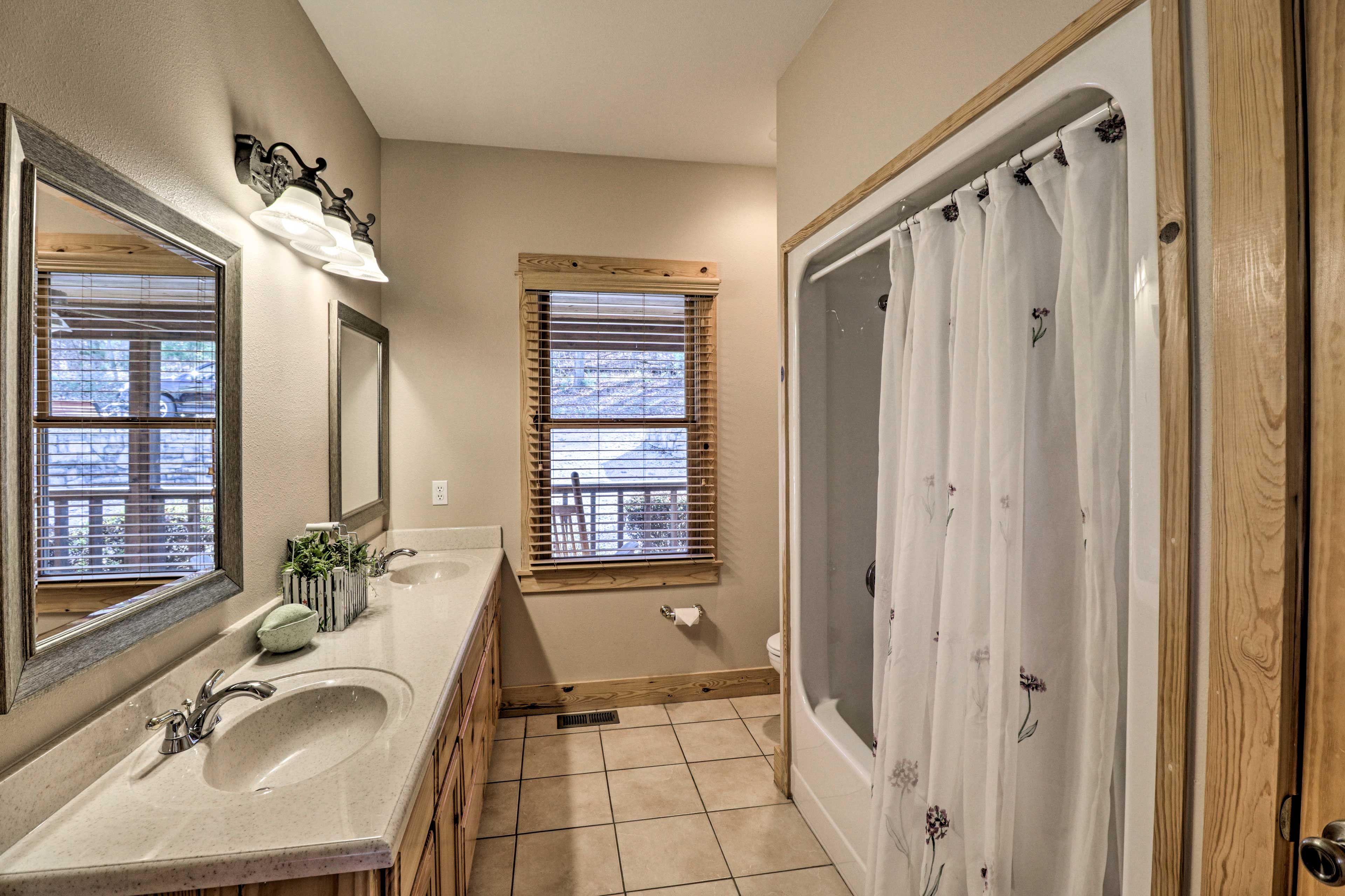 Une salle de bain avec un grand lavabo, deux fenêtres équipées de stores, une douche avec un rideau à motifs floraux et un miroir avec un luminaire.