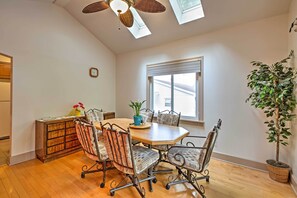 A dining area with a wooden table, four chairs, a clock, a window with blinds, and a potted plant.
