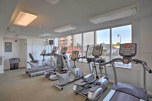 A gym with multiple treadmills and elliptical machines, large windows with a view of buildings, and a door with a sign. - Oceanfront Corner Condo in North Myrtle Beach! (North Myrtle Beach)