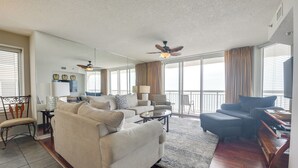 A spacious living room with a large sofa, a coffee table, and a ceiling fan. There is a dining area with a table and chairs, and a view of the outside through large windows.