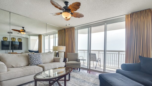 A spacious living room with a large sofa, a smaller sofa, a coffee table, and a ceiling fan. There is a large window with curtains and a view of the outdoors.