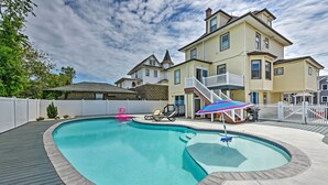 A swimming pool with a wooden deck, a white fence, and a house with a balcony in the background. - Seaside Wildwood Haven: Walk to Boardwalk/beach! (Wildwood Crest)