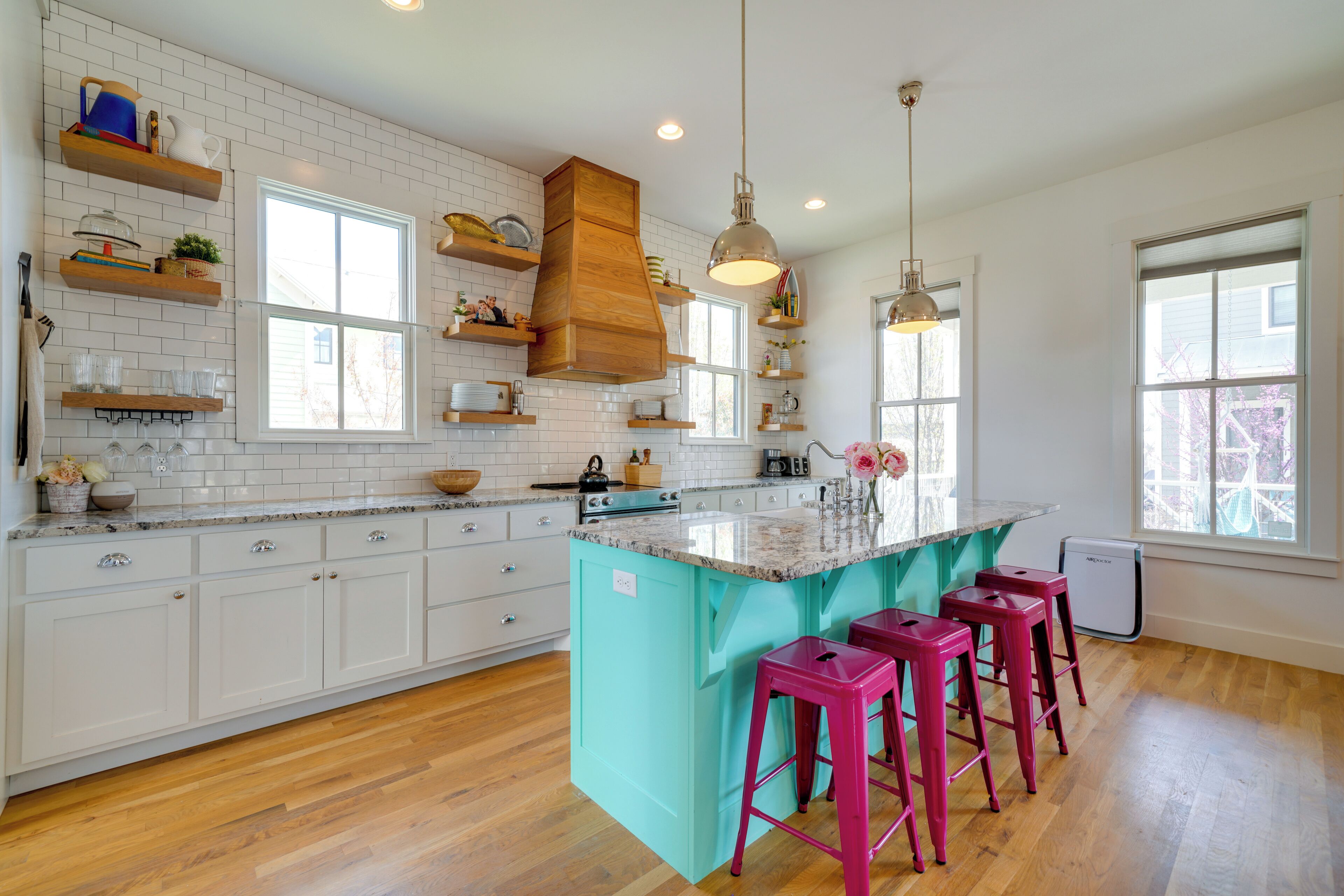 A kitchen with white cabinets, a turquoise island, wooden shelves, and pink bar stools.